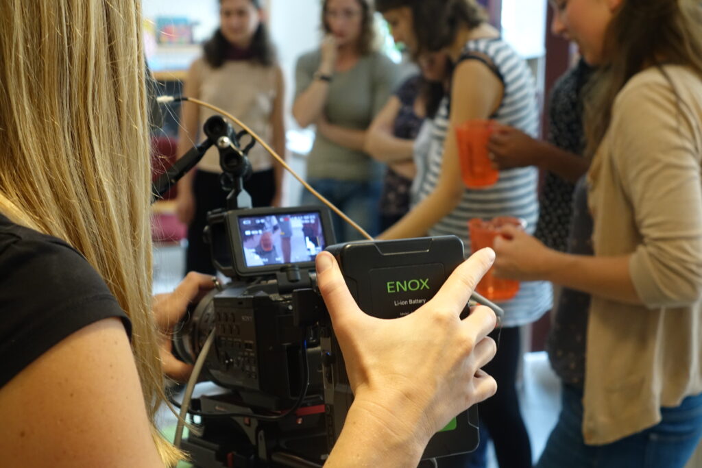 Frau im Vordergund mit Videokamera, im Hintergrund unscharf eine Gruppe von Jugendlichen.